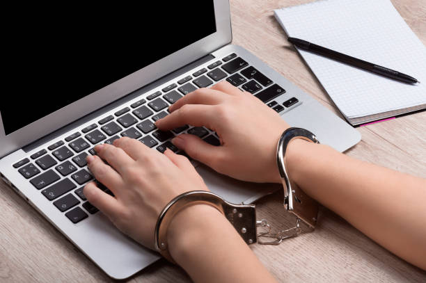 A girl in handcuffs is sitting at a laptop. The concept of restricting freedom of speech or punishing the disclosure of confidential information or the dissemination of fakes. Very close-up.  oppress stock pictures, royalty-free photos & images